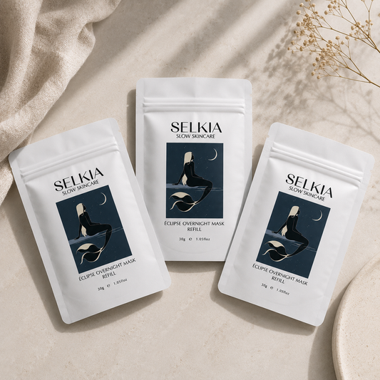 Three white stand-up pouches of SELKIA Slow Skincare Eclipse Overnight Mask Refill are arranged on a light beige surface with soft shadows, a draped fabric in the upper left, and dried floral stems in the upper right.