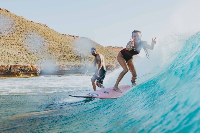 Two people surfing on a wave with a desert landscape in the background